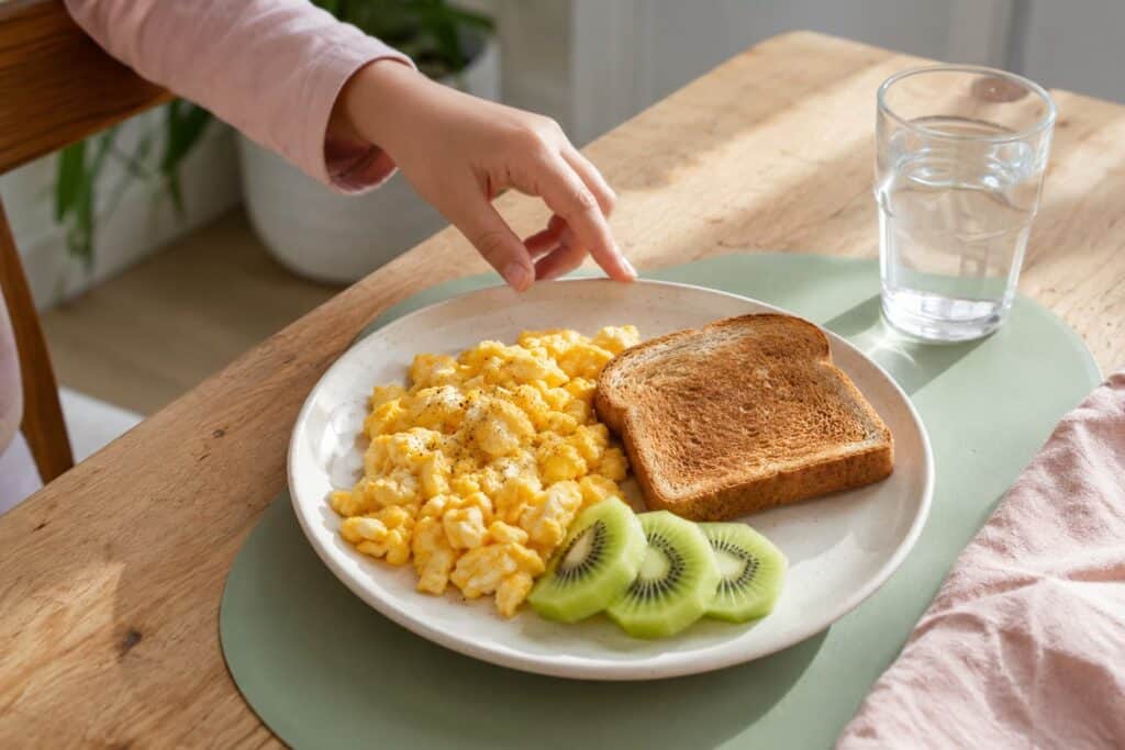 Child reaching for scrambled eggs with toast, kiwi and water at breakfast